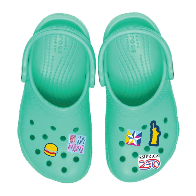 America250™ Shoe Charm-5 Pack Set displayed on mint green foam clogs with five colorful shoe charms, including patriotic and graphic designs, attached to ventilated slip-on footwear for shoe customization and personal style.