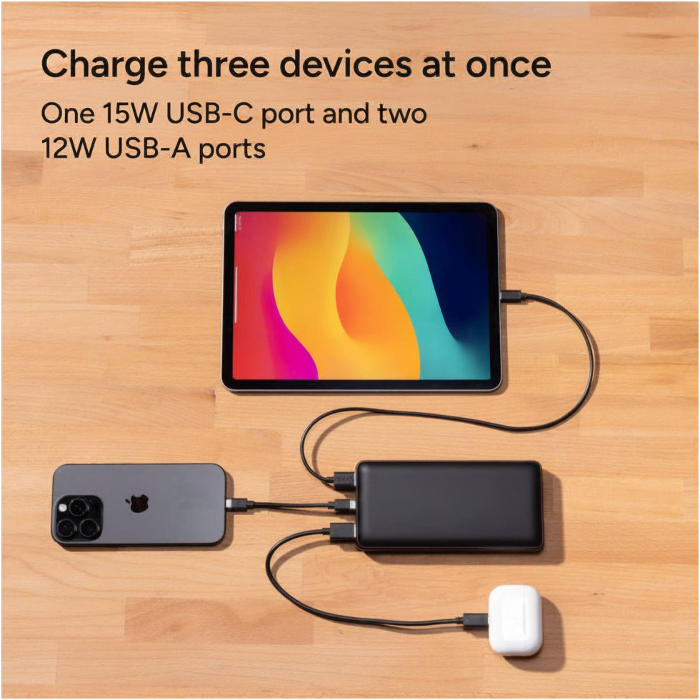 mophie Power Boost Pro XL black 20000mAh power bank shown on a table next to a smartphone and tablet with multiple devices connected, LED battery indicator visible while charging in a casual indoor home or office scene.