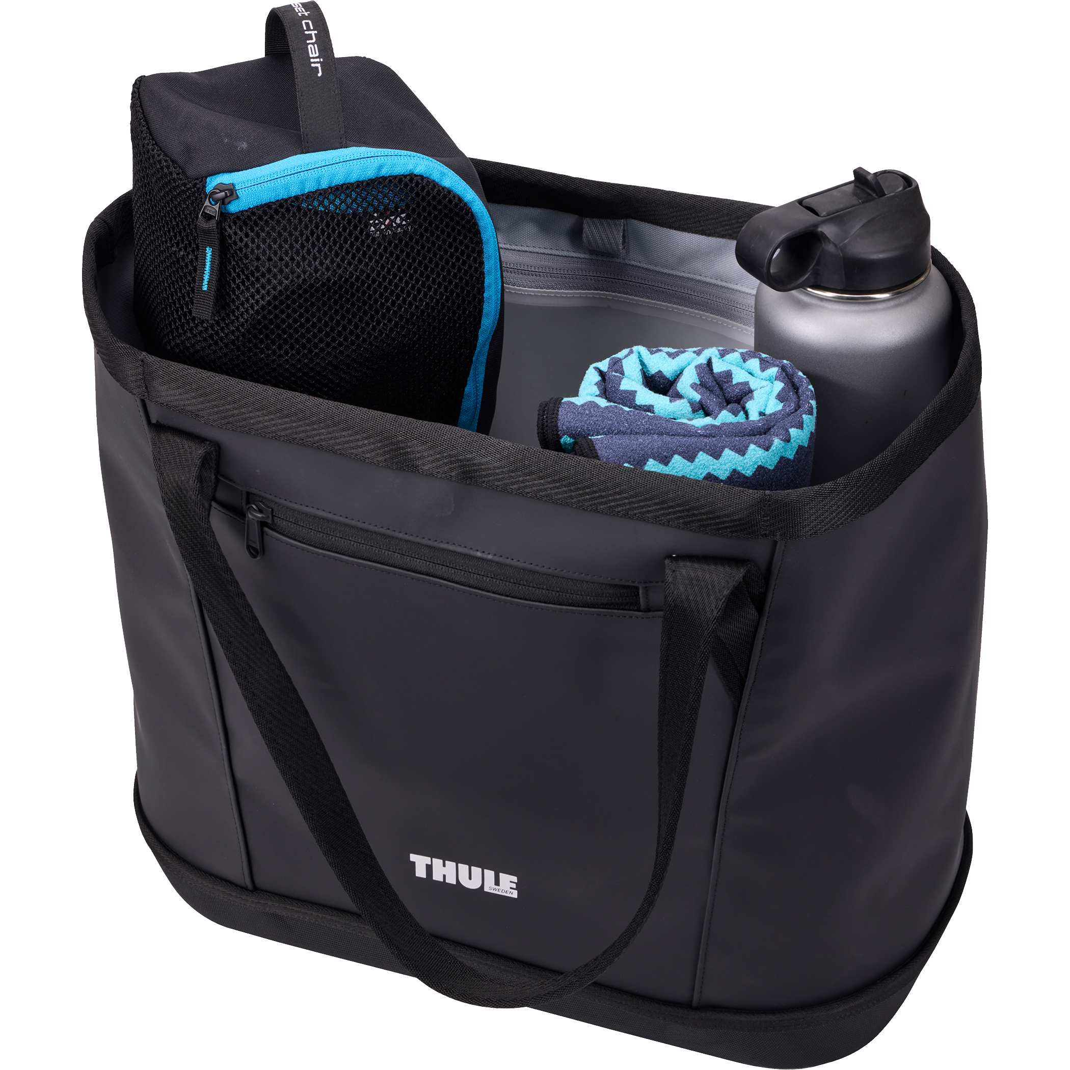 Thule 30L Chasm Gear Tote in black durable fabric with carry handles, placed outdoors on a rocky surface beside hiking and travel gear, showcasing everyday carry and adventure use in an outdoor setting.