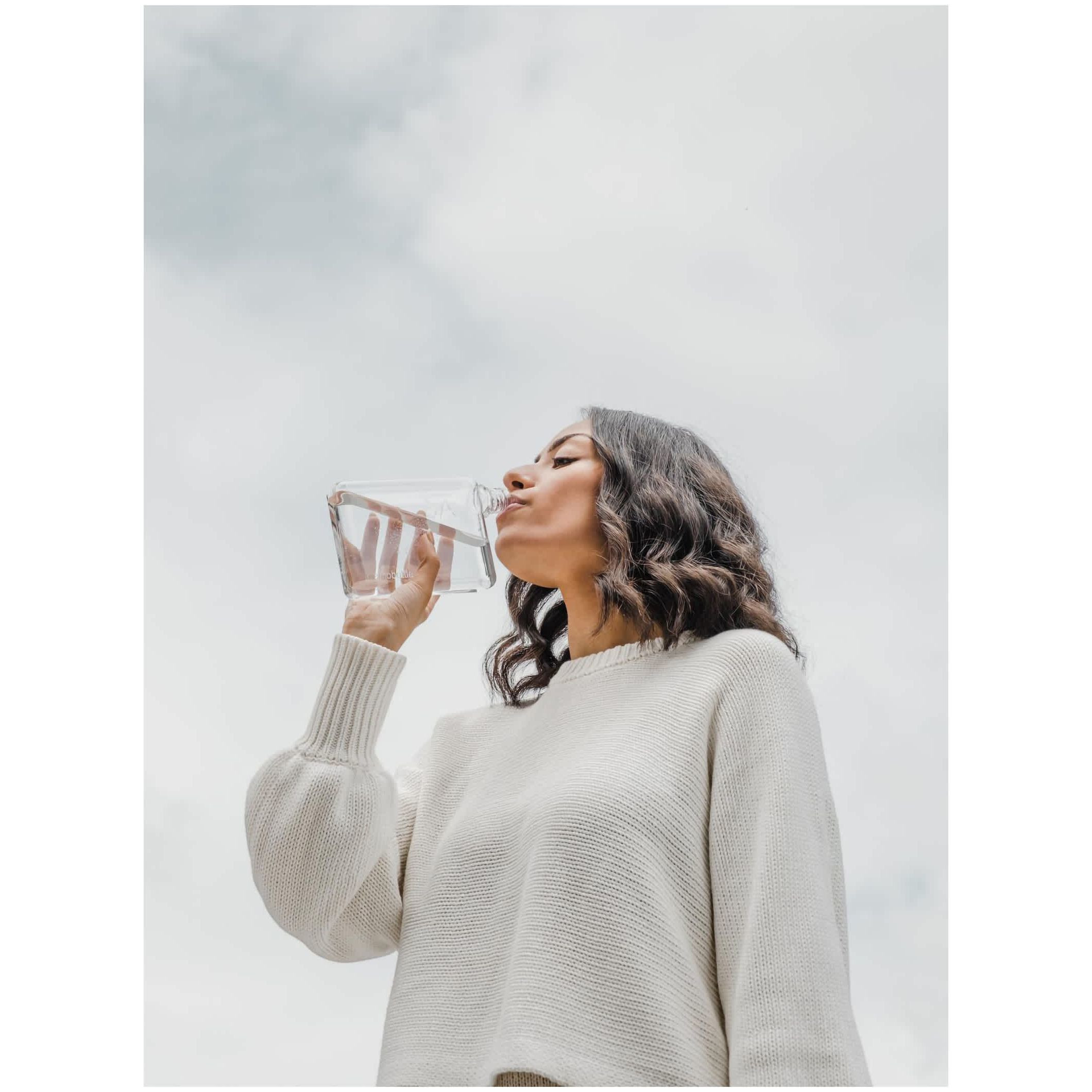 A6 Tritan™ memobottle™ in clear Tritan plastic with a stainless steel lid, held up to drink in an outdoor lifestyle setting, highlighting a slim reusable water bottle for everyday hydration.