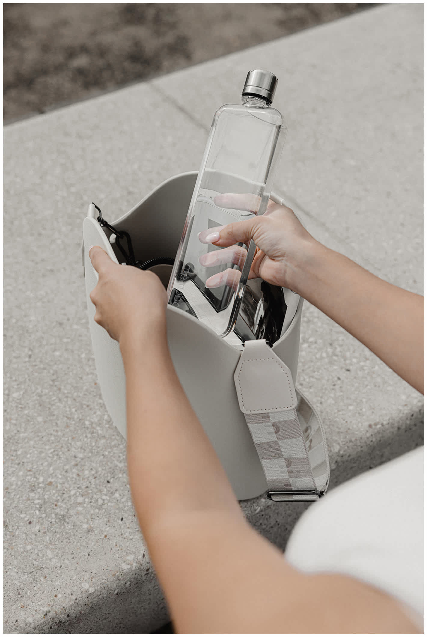 Clear Slim Tritan™ memobottle™ 15 oz. Tritan plastic water bottle with a stainless steel lid being placed into a light gray shoulder bag, showcasing its slim rectangular design for portable everyday hydration.