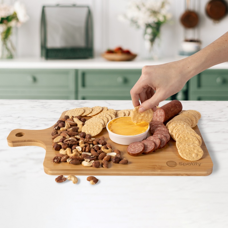 Wood bamboo charcuterie board with cheddar cheese spread, summer sausage, mixed salted nuts, and savory rice thin crackers, featuring a convenient handle for transport.