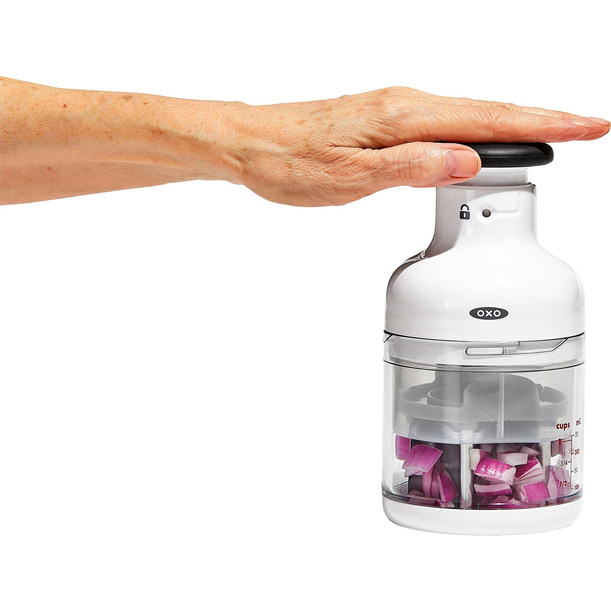 White OXO Good Grips Chopper, white plastic food chopper with a clear measurement-marked catcher cup and pump-action knob, featuring a rotating stainless steel blade for chopping vegetables, herbs, nuts, and other ingredients.